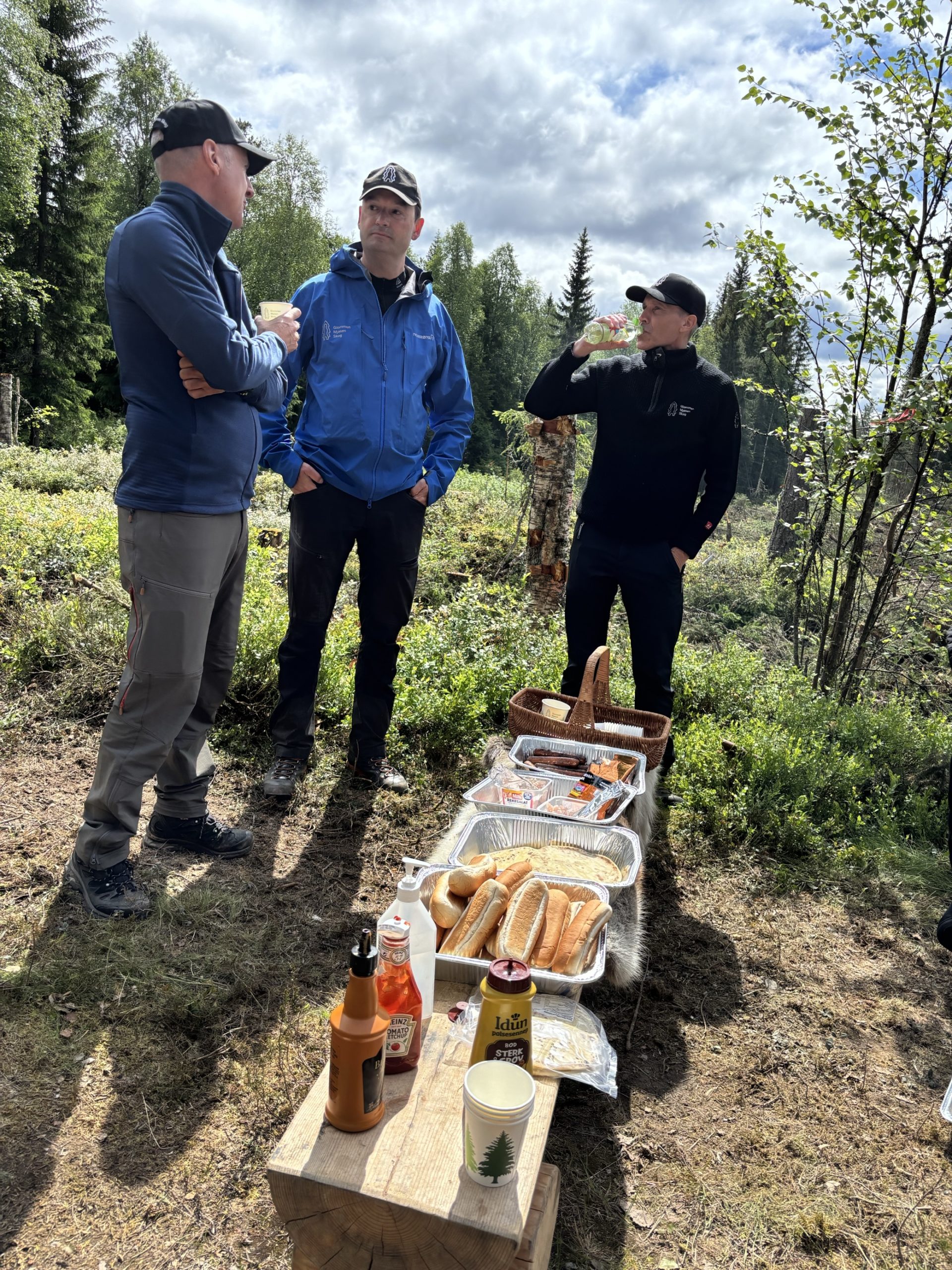 Tre menn ved benk med grillede pølser i skogen.